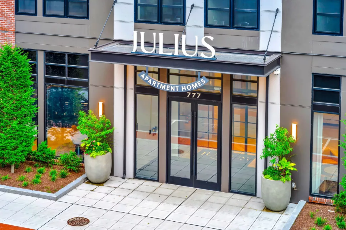 Entrance to an apartment community with a sign that says "Julius" over glass doors that are flanked by potted plants.
