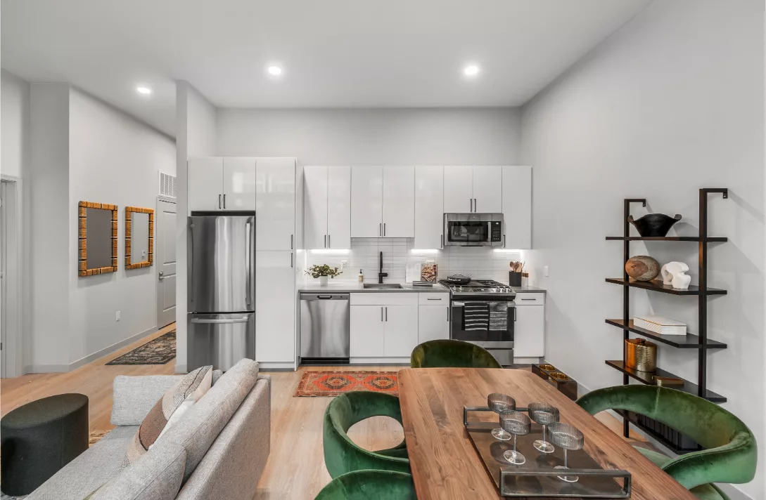 Open-plan living space with modern kitchen, stainless steel appliances, green velvet chairs, and a wood dining table.