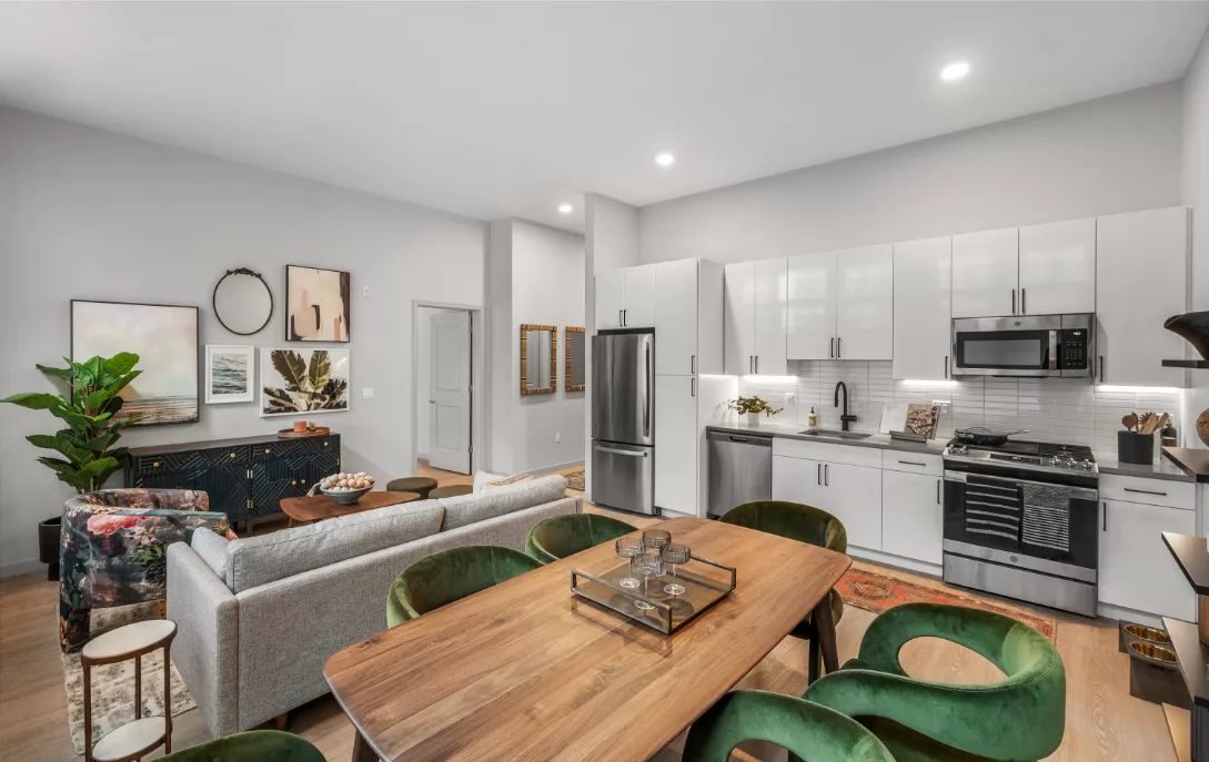 Open-plan apartment with kitchen with white cabinets and tile backsplash, dining area with wooden table and green velvet chairs, and living area with couch, armchairs, and modern artwork.